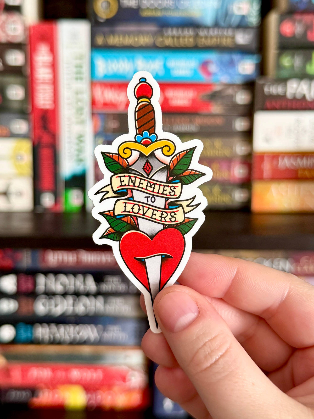 Sticker with a heart and sword design held in front of a bookshelf.