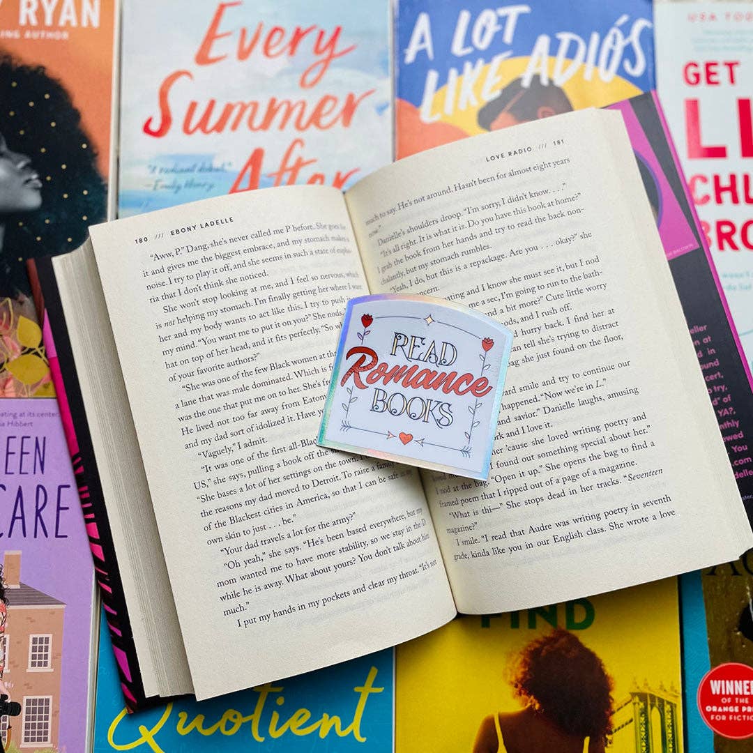 An open book spread with printed text, layered over several colorful books and a small square sticker that reads “Read Romance Books.” The scene is styled as a flat lay with no person or real environment visible.