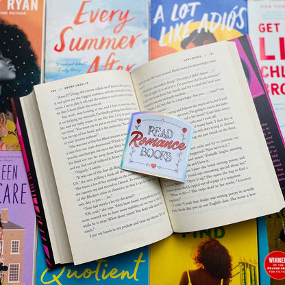 An open book spread with printed text, layered over several colorful books and a small square sticker that reads “Read Romance Books.” The scene is styled as a flat lay with no person or real environment visible.