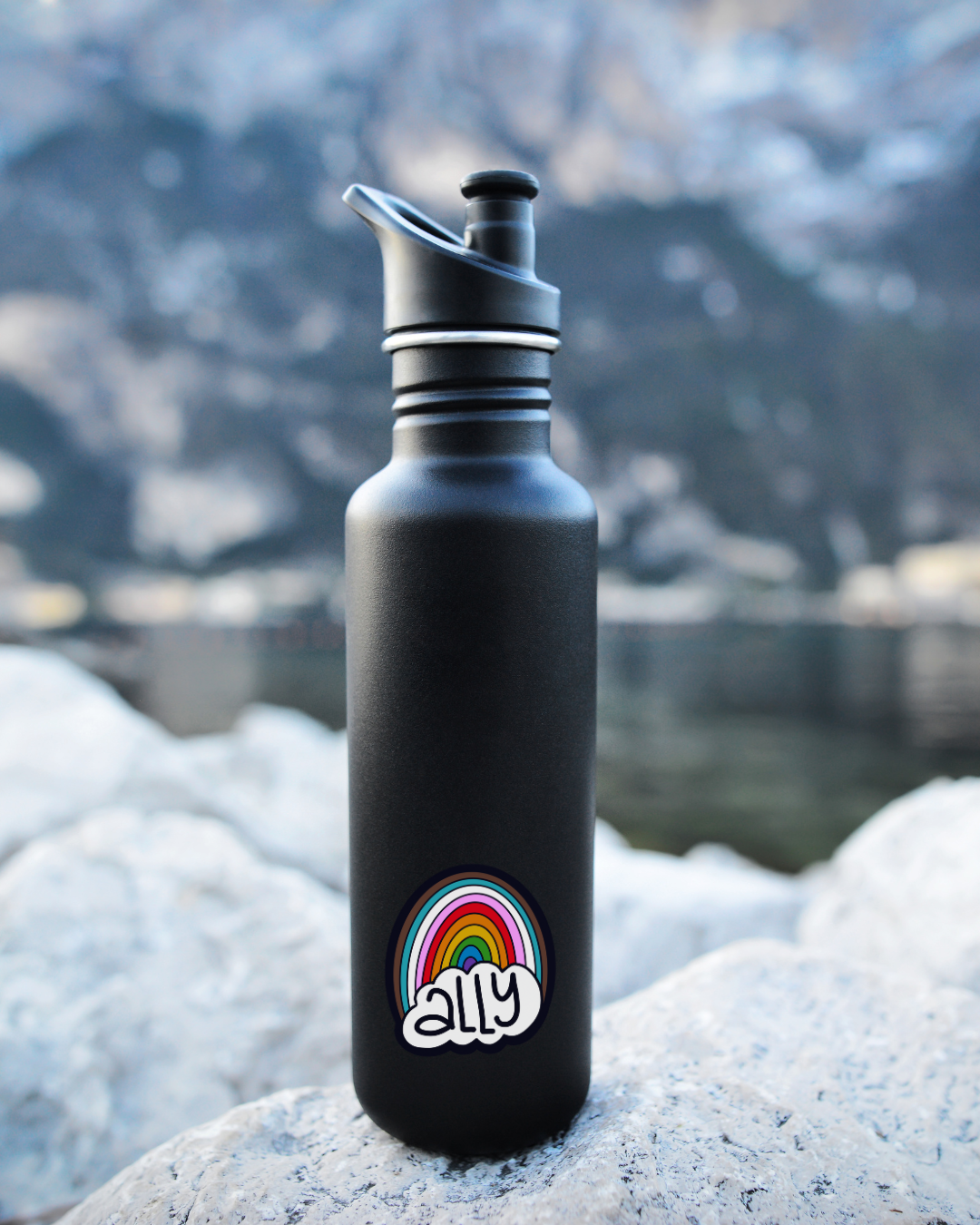Black water bottle with a colorful rainbow sticker on a rock with a mountainous background