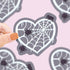 Heart-shaped spider web sticker held by a hand on a pink background with more stickers.