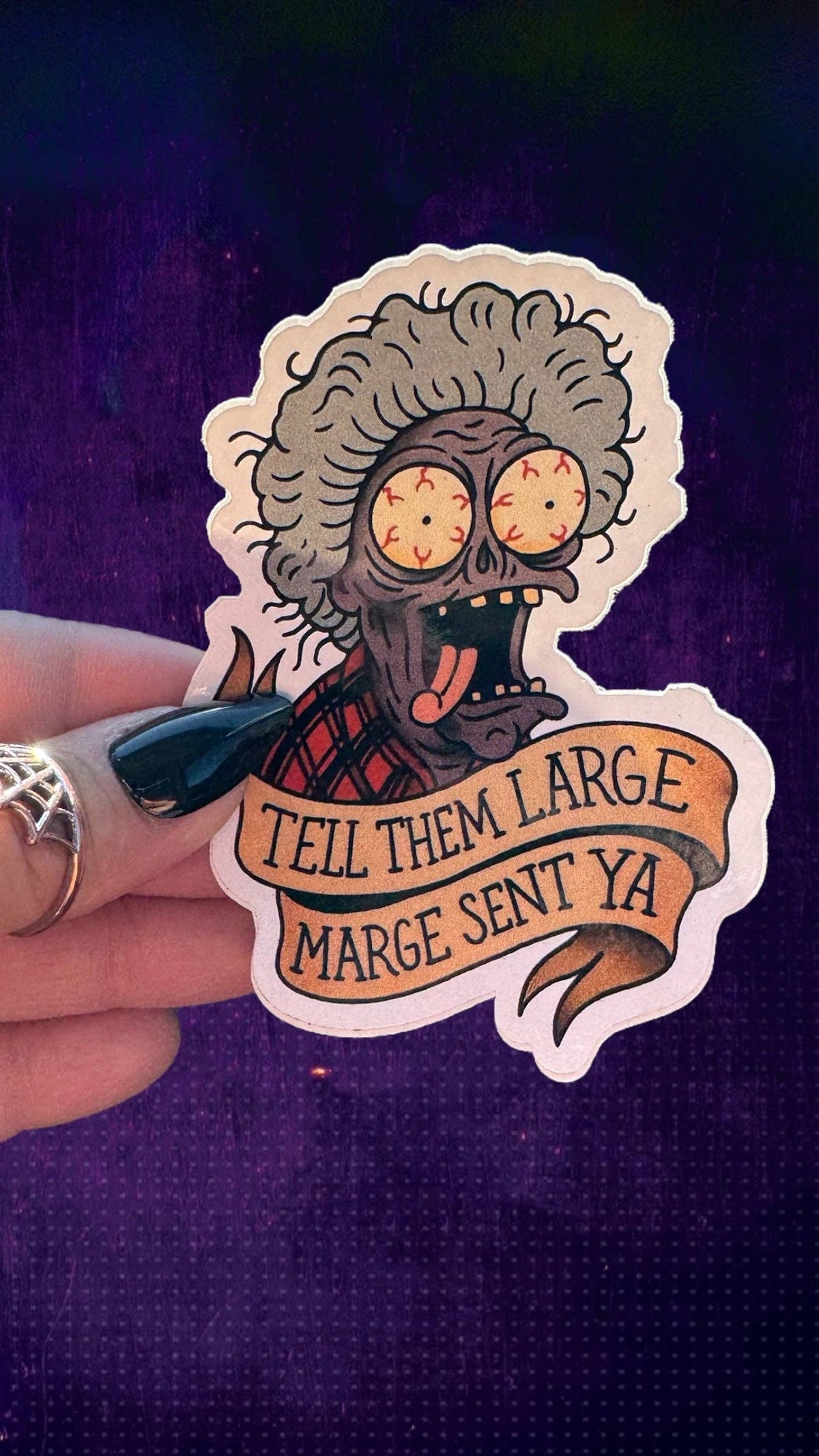 A hand holds a die-cut sticker featuring a cartoon zombie with gray hair, bulging yellow eyes, and a red plaid shirt. The sticker includes a banner reading &