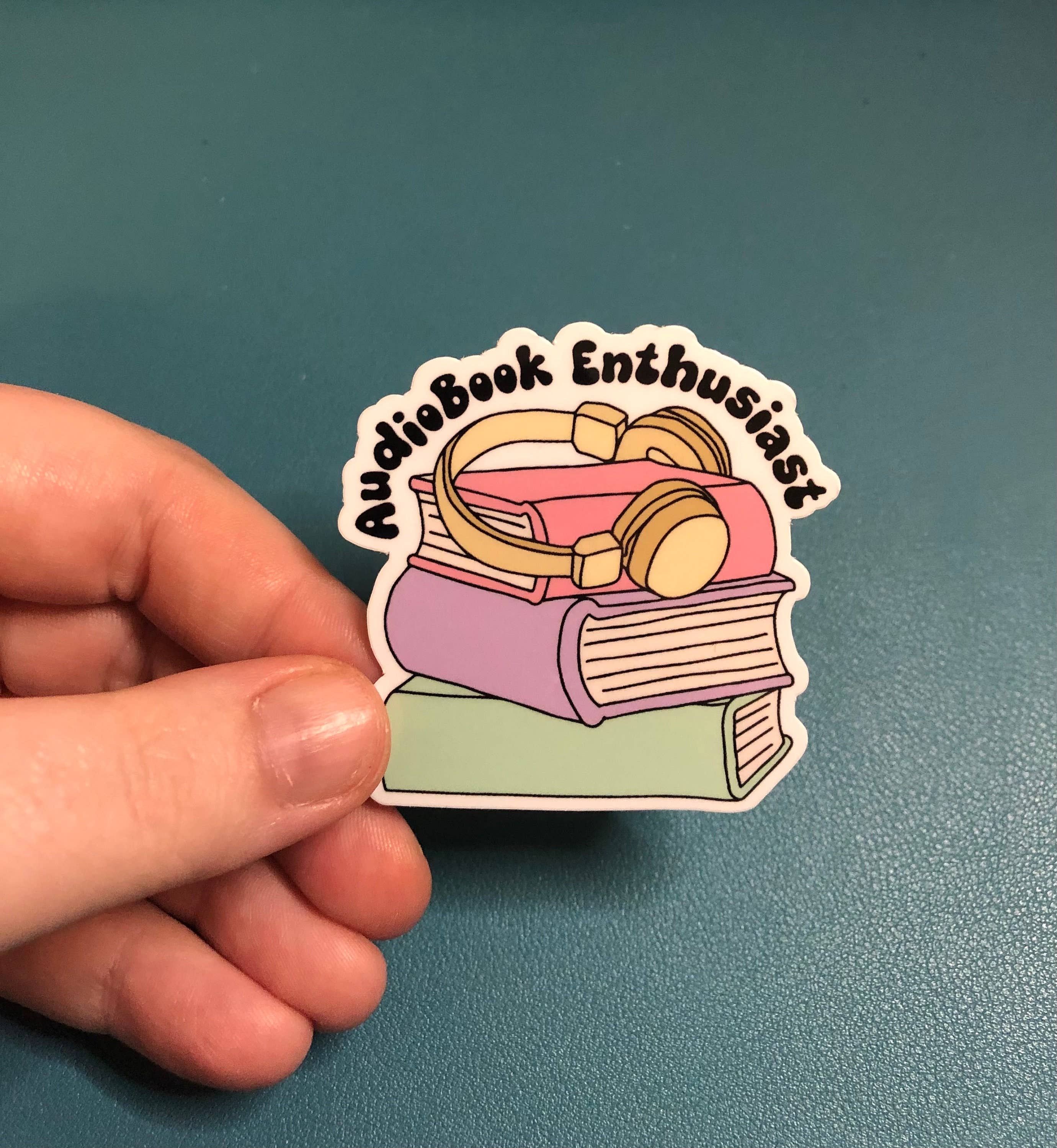 A hand holds a die-cut sticker featuring a stack of books with over-ear headphones on top and the words &