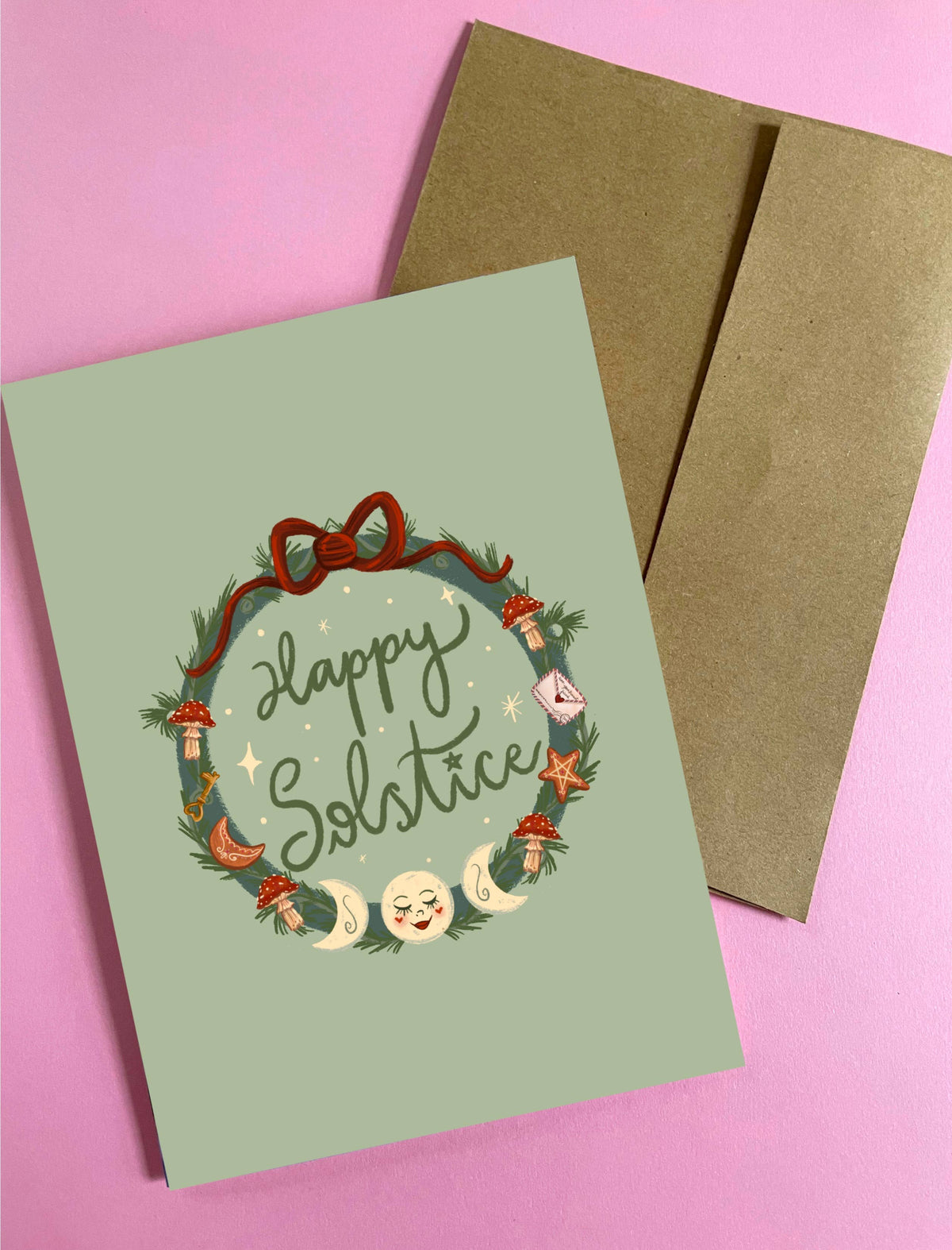 A square greeting card with a sage green background and a festive wreath illustration reading &