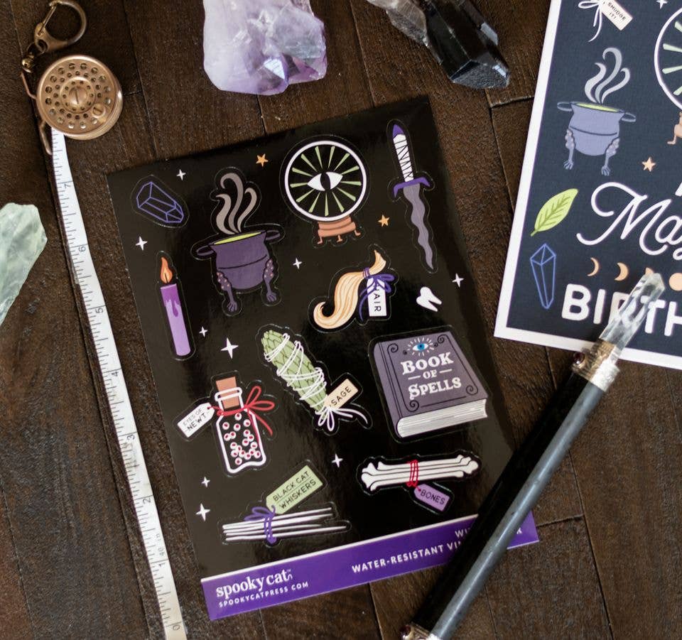 A sheet of water-resistant vinyl stickers featuring spooky witch and potion illustrations, displayed on a dark wooden tabletop with crystals, a compass, and a card.
