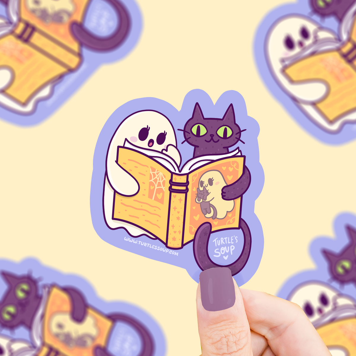 A hand with purple nail polish holds a die-cut sticker featuring a white ghost and a black cat reading a yellow book. The sticker is shown against a pale yellow background with blurred repeating versions of the same sticker design.