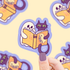 A hand with purple nail polish holds a die-cut sticker featuring a white ghost and a black cat reading a yellow book. The sticker is shown against a pale yellow background with blurred repeating versions of the same sticker design.