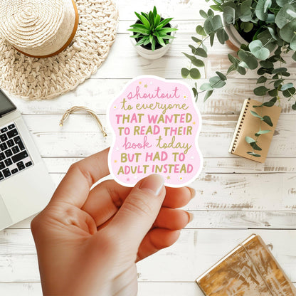 Hand holding a pink sticker with text on a desk with laptop, notebook, and plants
