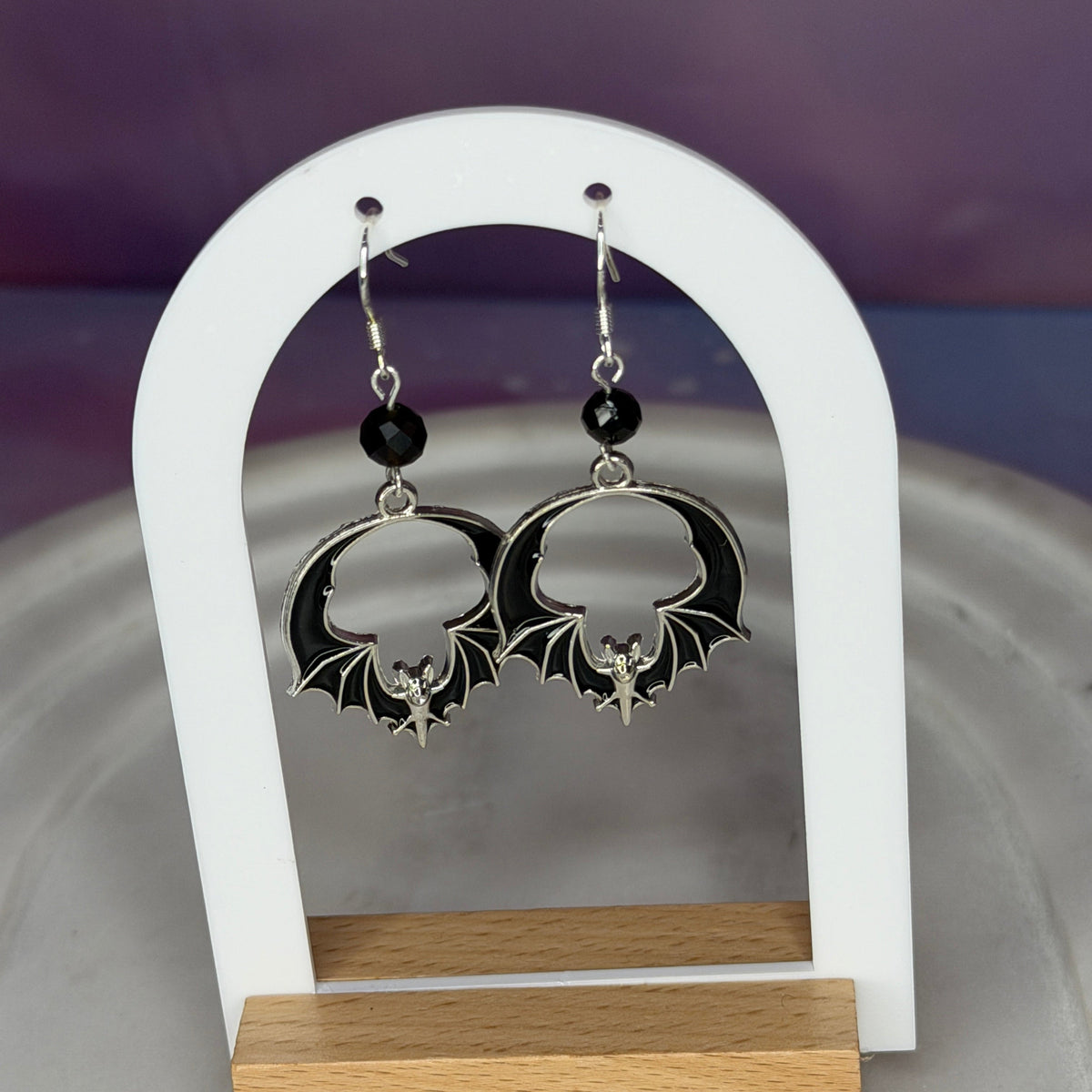 A pair of gothic-style dangle earrings displayed on a white arched stand with a wooden base. The earrings feature silver bat-shaped charms with black wings, a small silver fairy figure, and a black bead above the bat charm.