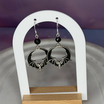 A pair of gothic-style dangle earrings displayed on a white arched stand with a wooden base. The earrings feature silver bat-shaped charms with black wings, a small silver fairy figure, and a black bead above the bat charm.