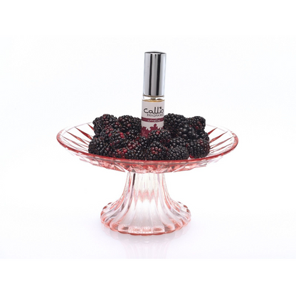 Image of a bottle of Amelia Travel-Size Perfume Spray by Callio Fragrance on a pink pedestal, surrounded by blackberries on a white background.