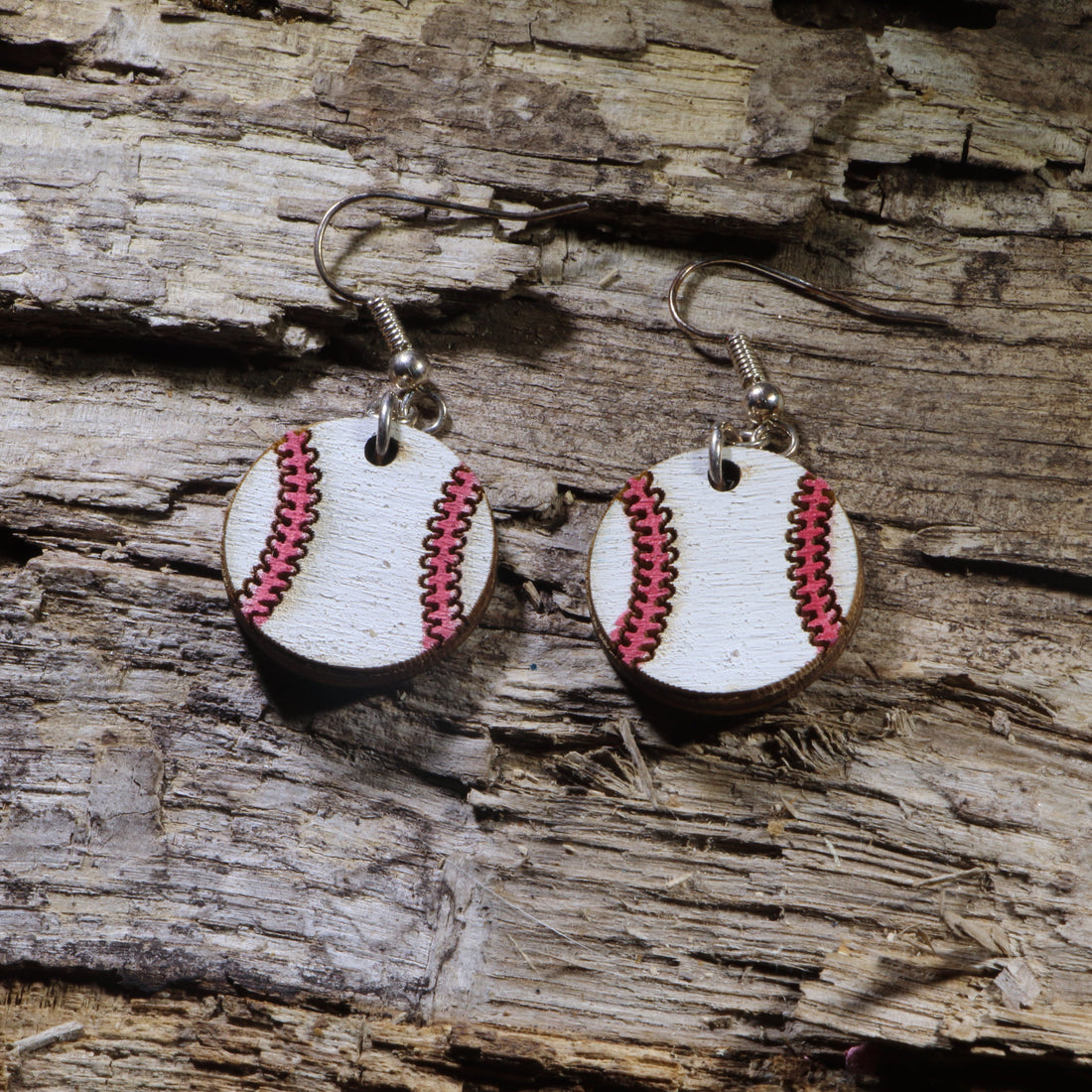 Handmade wooden baseball dangle earrings featuring white baseballs with pink stitched seams, hanging from silver-tone hypoallergenic hooks on a rustic wood background.
