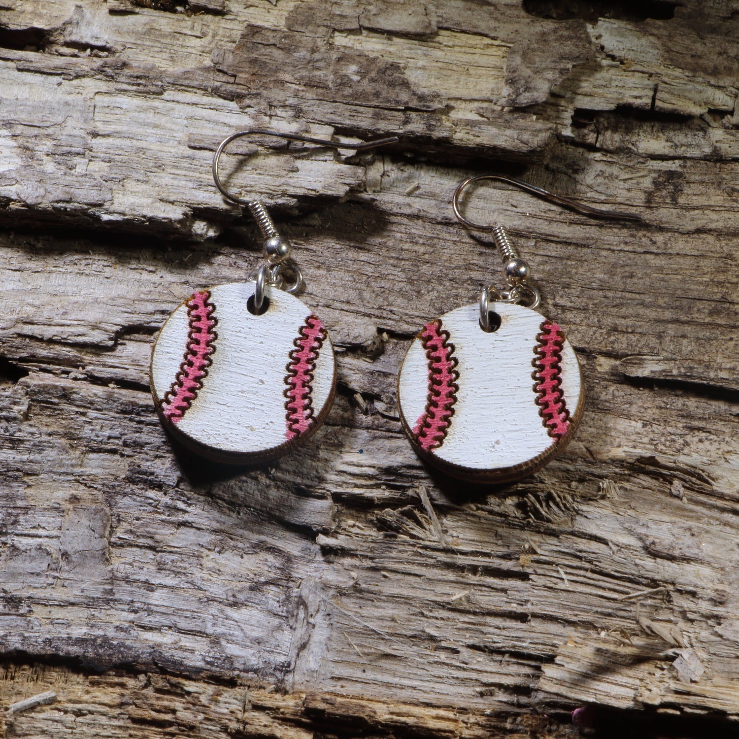 Handmade wooden baseball dangle earrings featuring white baseballs with pink stitched seams, hanging from silver-tone hypoallergenic hooks on a rustic wood background.
