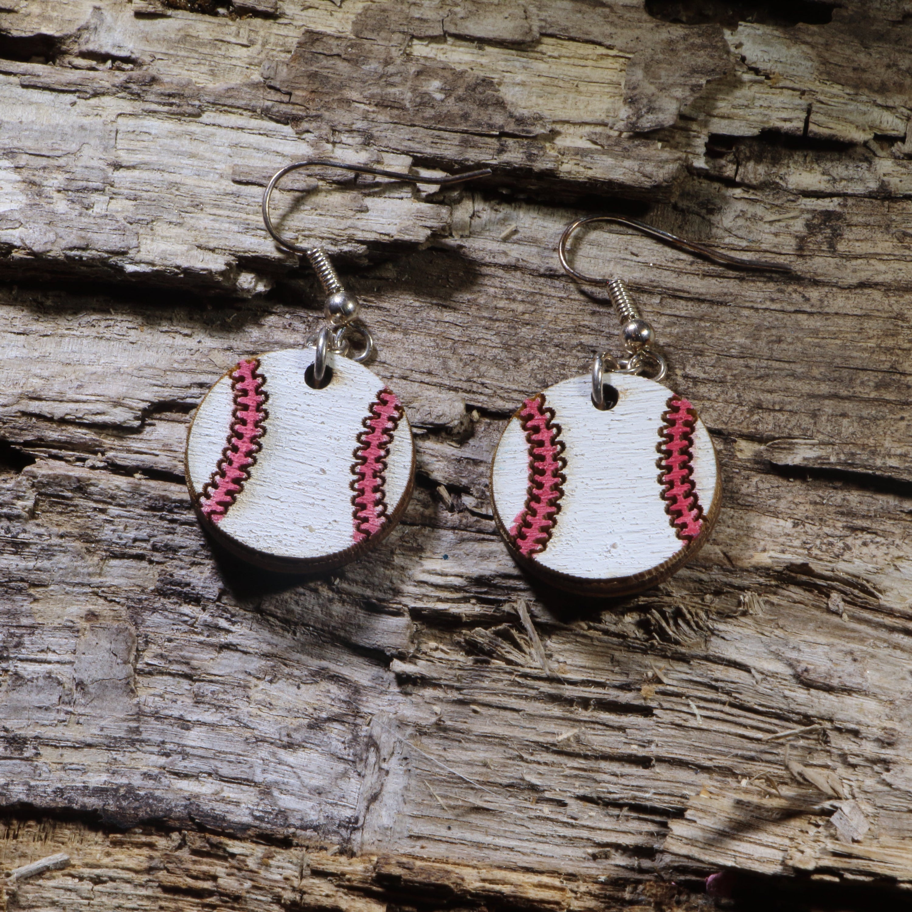 Handmade wooden baseball dangle earrings featuring white baseballs with pink stitched seams, hanging from silver-tone hypoallergenic hooks on a rustic wood background.
