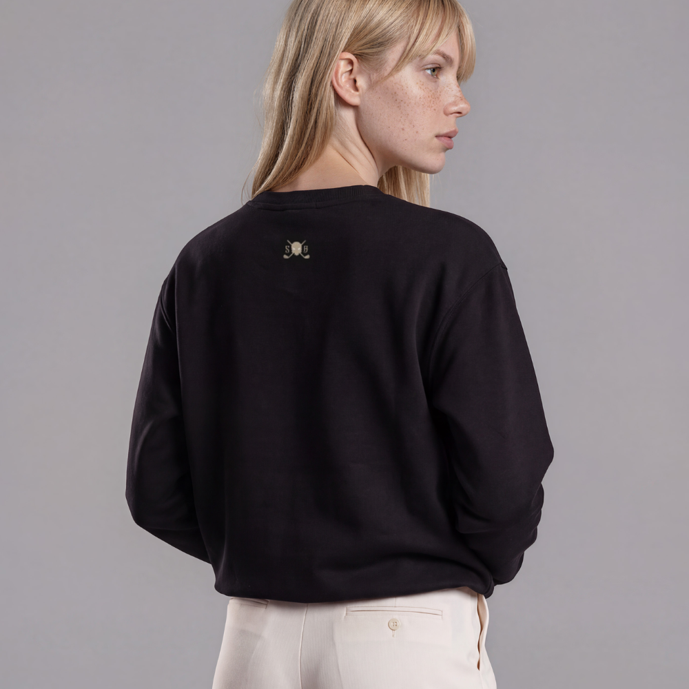 A woman shown from the back wearing a black crewneck sweatshirt with a small gray Skull &amp; Bogeys Golf Co. logo on the upper back, paired with cream-colored wide-leg trousers, against a solid gray studio background.