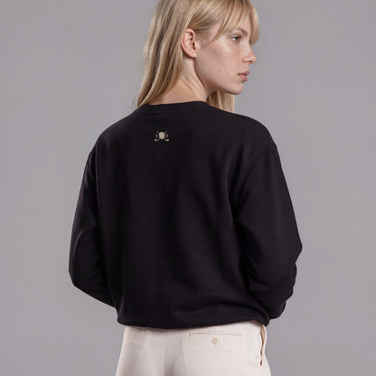 A woman shown from the back wearing a black crewneck sweatshirt with a small gray Skull &amp; Bogeys Golf Co. logo on the upper back, paired with cream-colored wide-leg trousers, against a solid gray studio background.