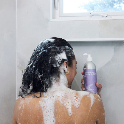 Person in a shower with soap suds, holding a natural citrus lavender body wash in an eco friendly aluminum bottle that can be returned to be refilled and reused. 