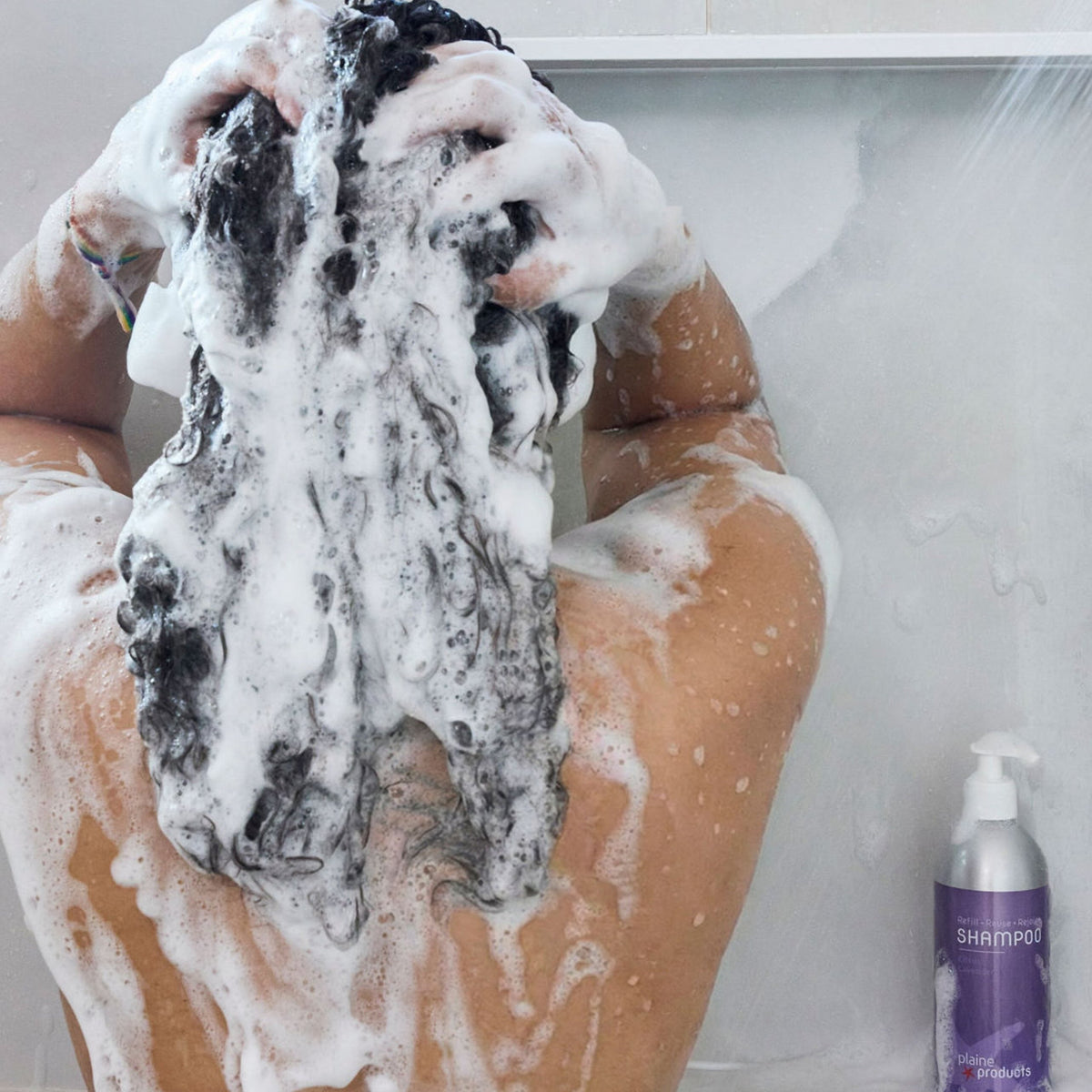 Person with lather and suds on the head and body, with a bottle of aloe-based, natural citrus lavender shampoo in the background.