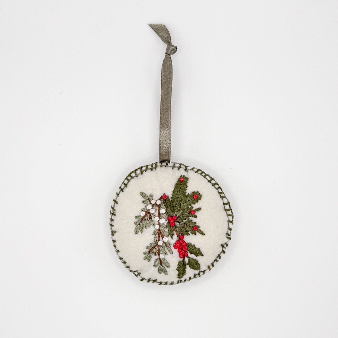 Ethically made by Syrian Refugees, this circular felt and hand embroidered ornament has a green ribbon, and red white and green cedar and berry motif, flat lay