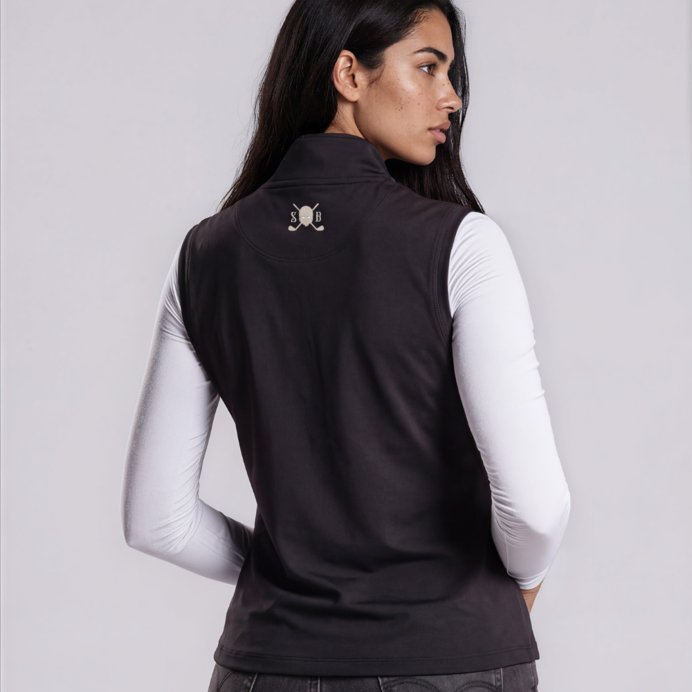 A woman shown from the back wearing a sleeveless black vest over a white long-sleeve top and dark gray jeans, against a light gray studio background.