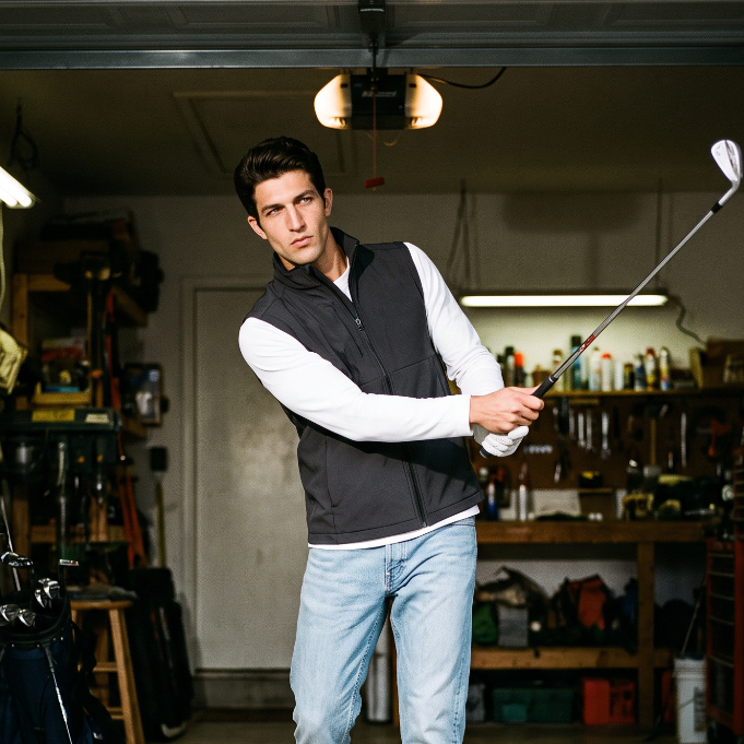 A man in a garage wearing a black zip-up vest over a white long-sleeve shirt, light blue jeans, and white sneakers, holding a golf club. The garage is filled with tools, equipment, and shelves.