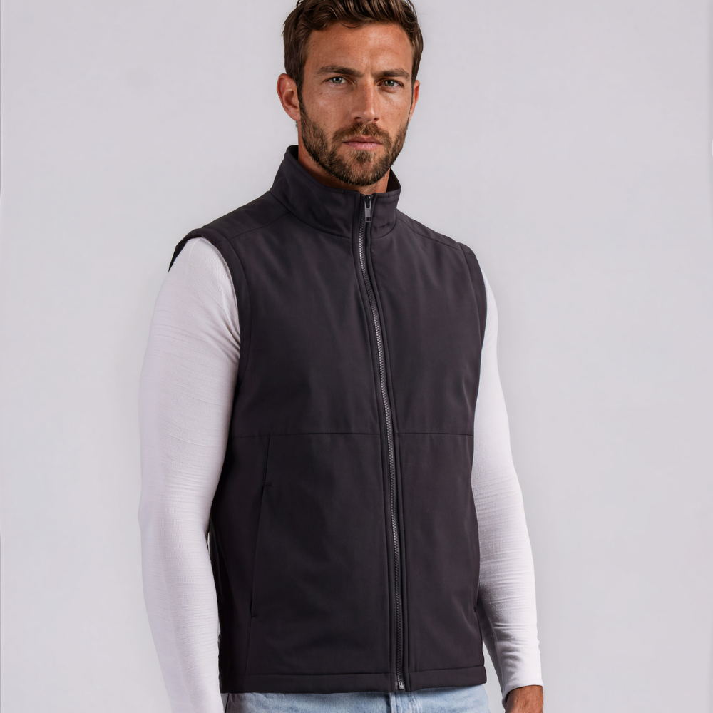 A man wearing a dark gray zip-up sleeveless vest over a white long-sleeve shirt and light blue jeans, standing against a light gray studio background.