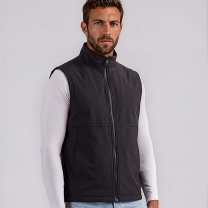 A man wearing a dark gray zip-up sleeveless vest over a white long-sleeve shirt and light blue jeans, standing against a light gray studio background.
