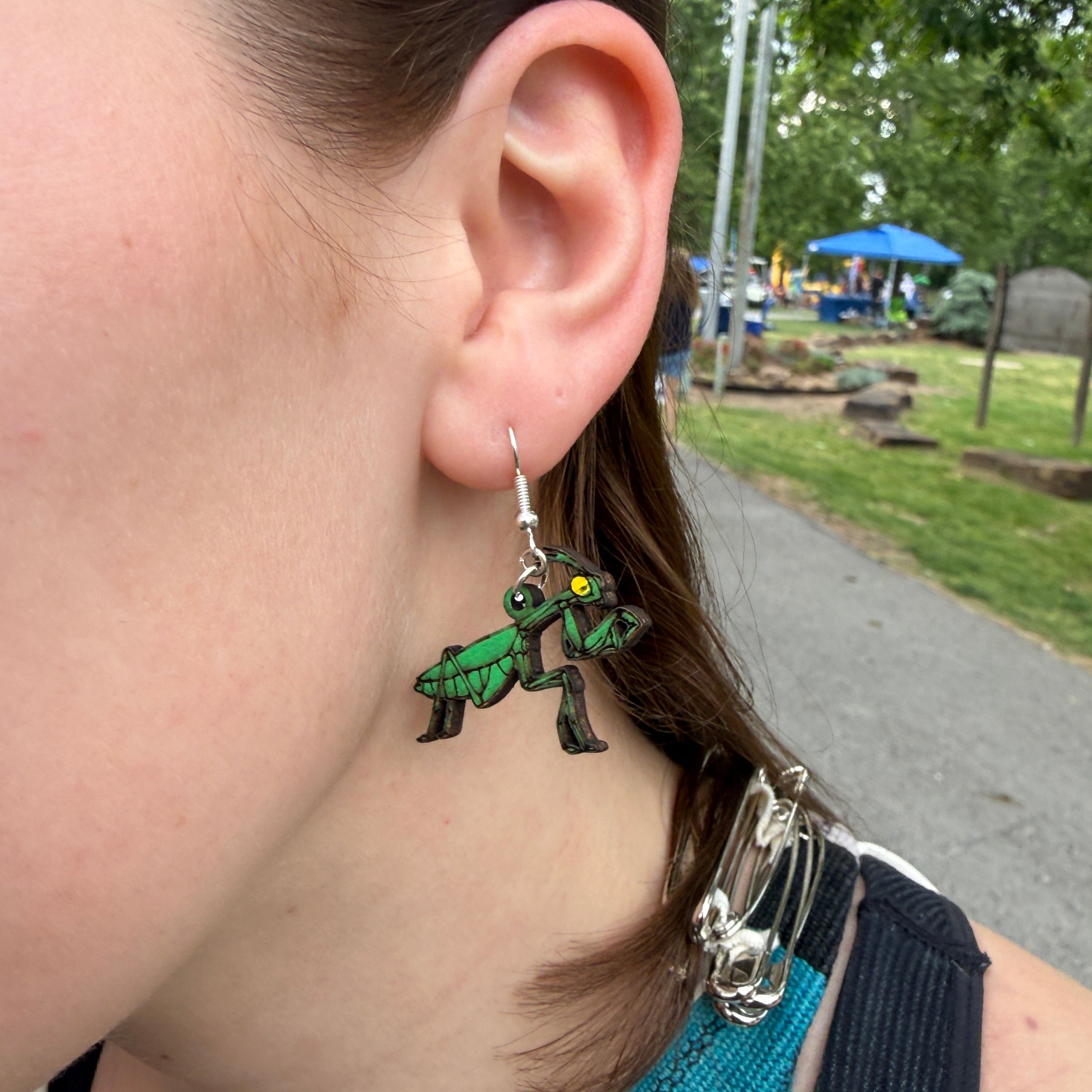 Close-up of a customer wearing hand-painted green praying mantis wooden dangle earrings. This lifestyle photo highlights the intricate detailing, lightweight design, and comfortable fit of these handmade insect earrings with hypoallergenic .925 sterling silver hooks. Perfect for insect enthusiasts, entomologists, gardeners, nature lovers, and anyone who enjoys unique bug-inspired jewelry.