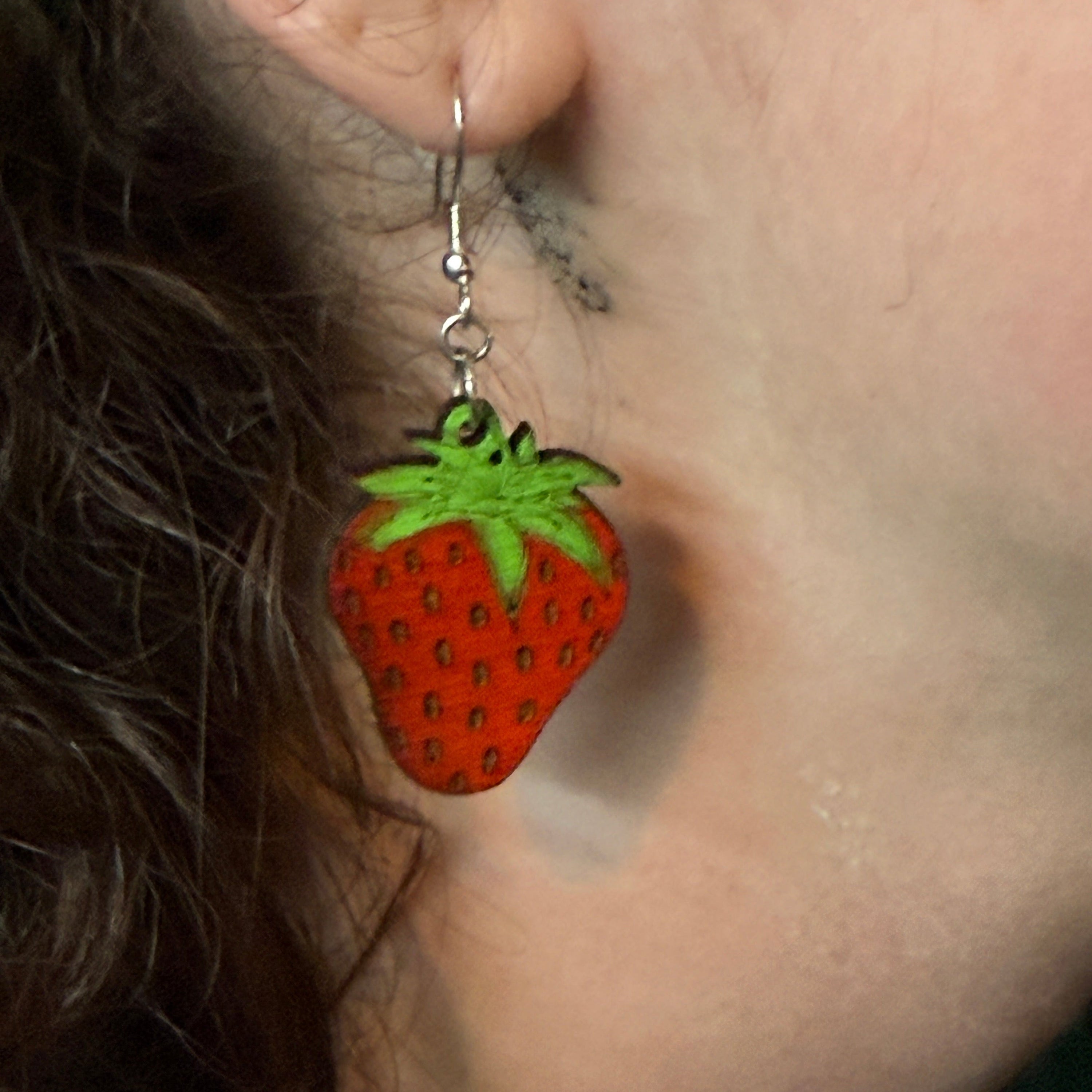 Close-up of a hand-painted wooden strawberry dangle earring featuring a realistic red strawberry with detailed seeds and bright green leaves. This detailed view showcases the handcrafted artwork and lightweight design with a hypoallergenic .925 sterling silver hook, perfect for fruit lovers, gardeners, summer accessories, and everyday statement jewelry.