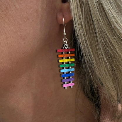Close-up of a person wearing hand-painted wooden rainbow xylophone dangle earrings on hypoallergenic .925 sterling silver hooks, highlighting the lightweight fit and vibrant rainbow colors.