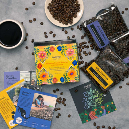 Overhead shot of a Coffee Tour box featuring women of Latin America; three coffee bags, with three info cards, a cup of coffee and beans scattered on a gray surface