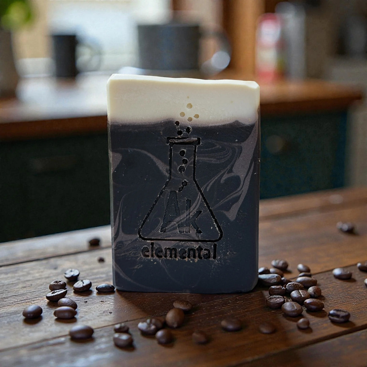 One brown soaps with creamy top sitting on a table with coffee beans surrounding the soap. AK Elemental logo of a erlenmeyer flask is stamped on the soap.