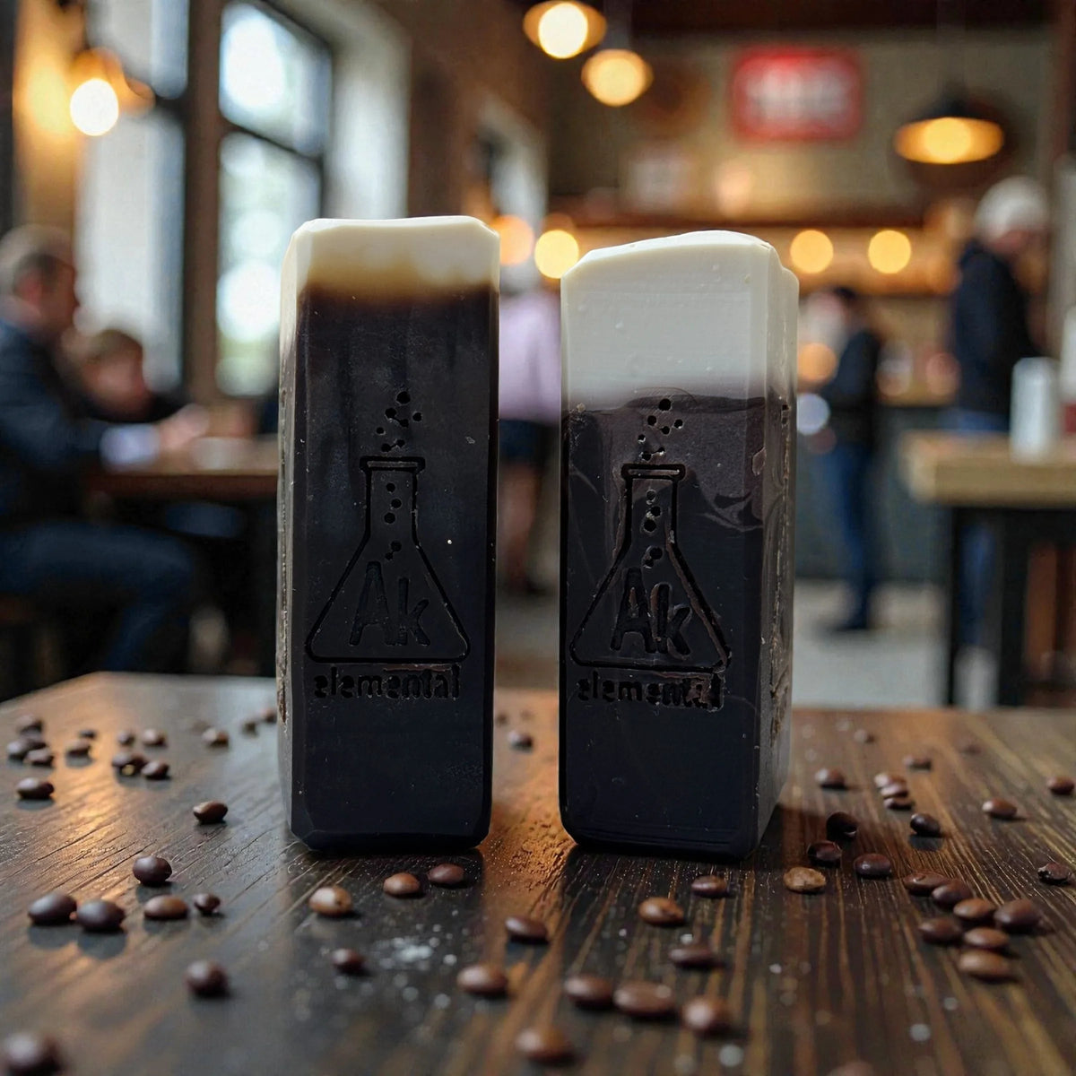 Side view of two brown soaps with creamy tops sitting on a table with coffee beans surrounding the soaps.