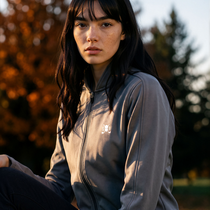 A woman wearing a gray zip-up hoodie with a small white embroidered logo on the left chest, photographed outdoors with trees and warm sunlight in the background.