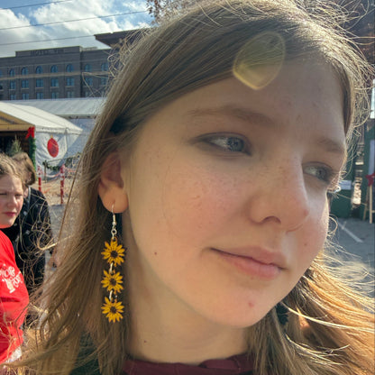 Customer wearing hand-painted cascading sunflower wooden dangle earrings featuring three vibrant yellow sunflowers with detailed brown centers. This lifestyle photo showcases the lightweight design, graceful movement, and comfortable fit of these handmade floral earrings with hypoallergenic .925 sterling silver hooks. Perfect for sunflower lovers, nature-inspired fashion, cottagecore outfits, and everyday wear.
