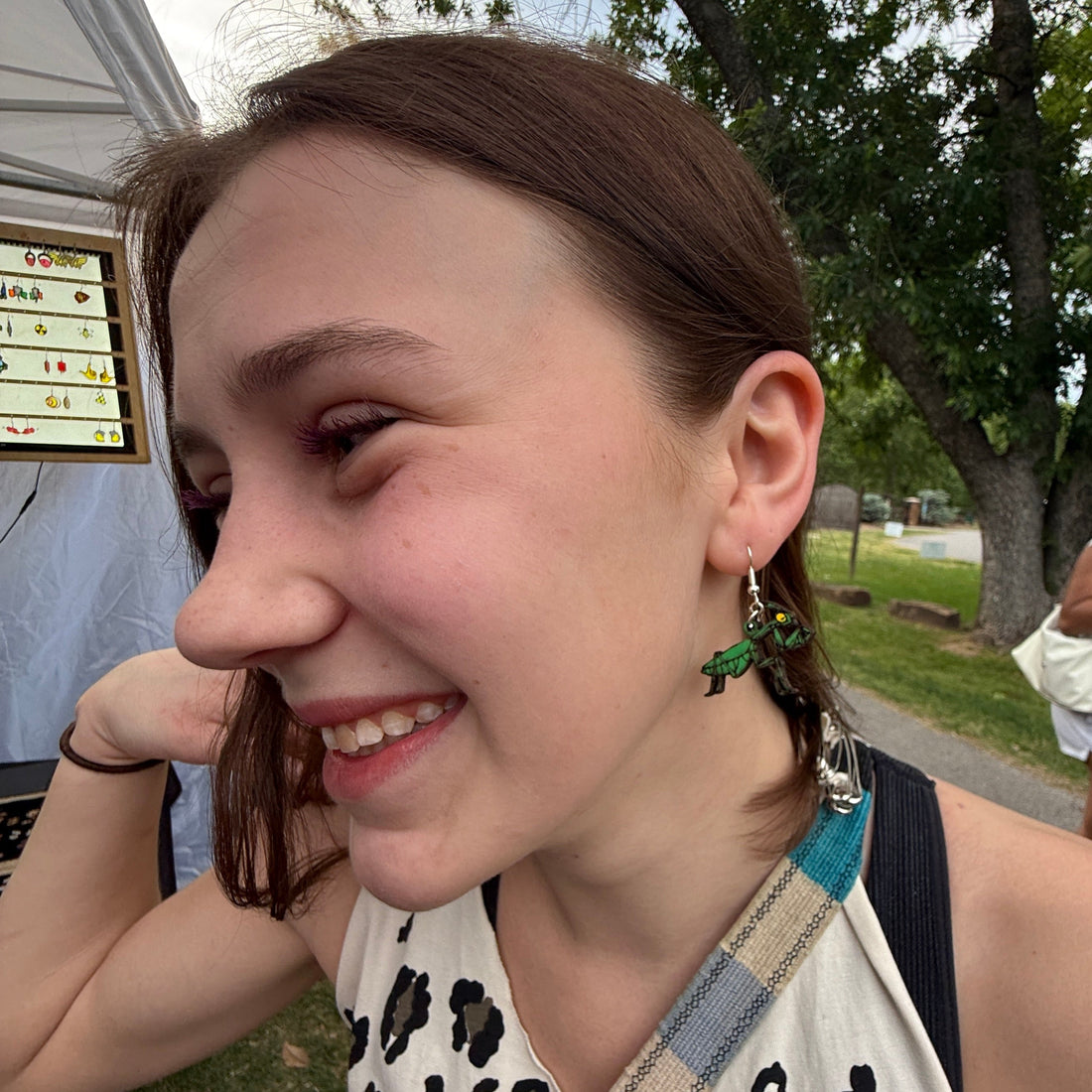 Customer wearing hand-painted green praying mantis wooden dangle earrings at an outdoor art festival. This lifestyle photo showcases the size, movement, and everyday wearability of these lightweight handmade insect earrings with hypoallergenic .925 sterling silver hooks. A distinctive accessory for bug lovers, nature enthusiasts, science teachers, and fans of unusual statement jewelry.