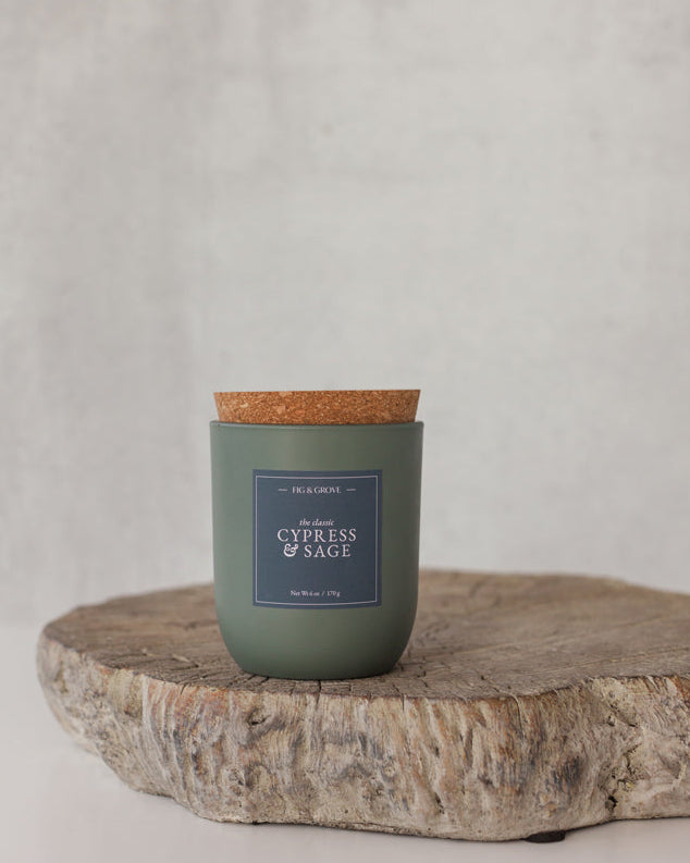 Cypress &amp; Sage Candle in a sage green glass jar with cork lid on a wooden surface against a light gray background