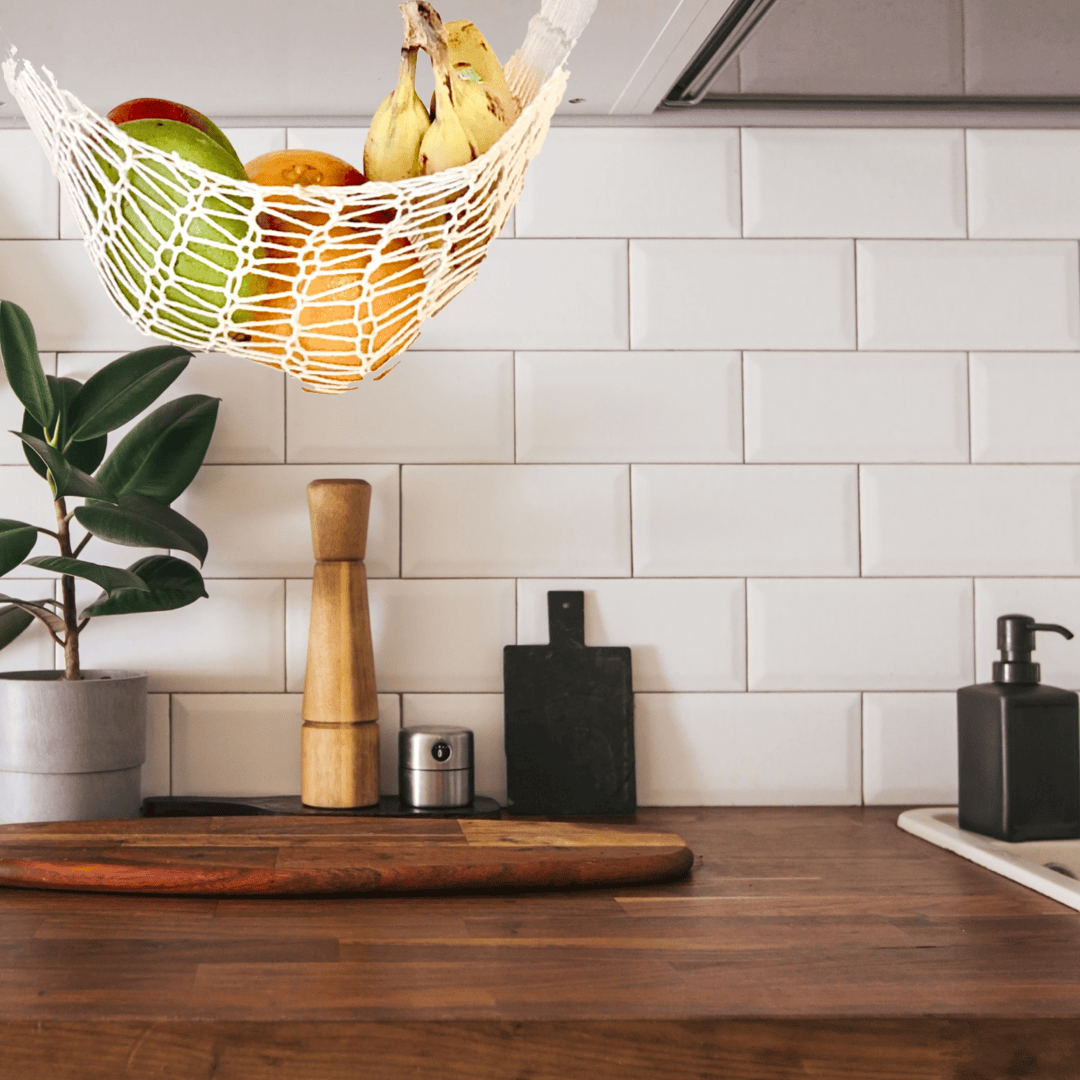 A Virginia-grown cotton produce hammock hangs from an upper counter, filled with bananas, oranges on mango. In the background is a subway tile backsplash. The counter is a dark wood, with a plant, wooden pepper grinder, small metal jar, metal bottle, and a metal pump bottle, and a wood cutting board on it.