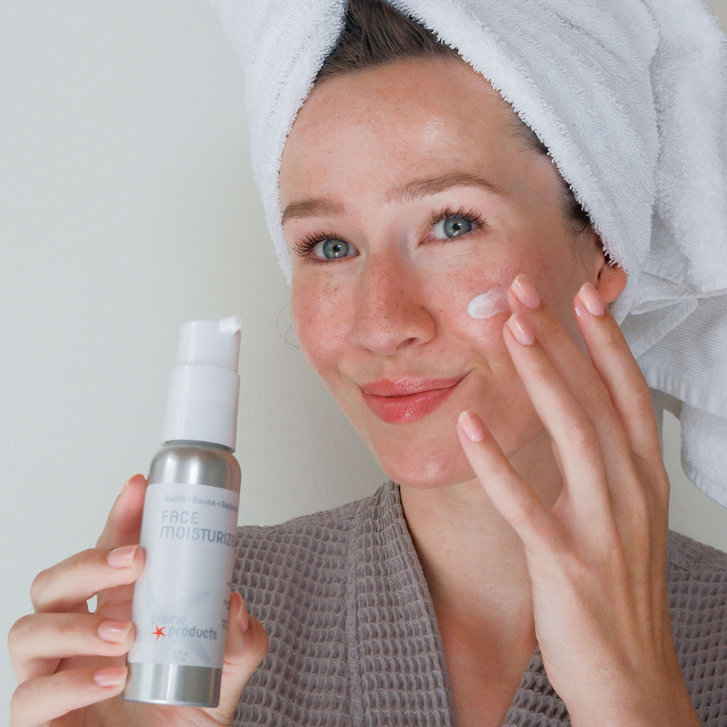 Woman applying eco friendly face moisturizer with a towel on her head, holding a plastic free bottle of skincare.
