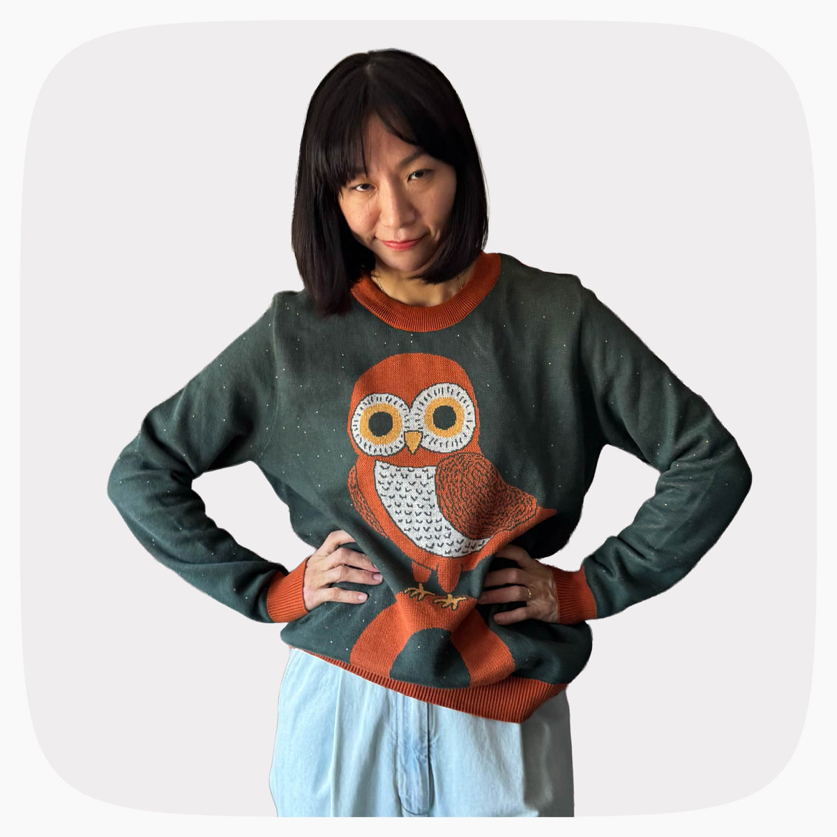 A woman wearing a green graphic knit sweater with an owl standing on the letter O design.