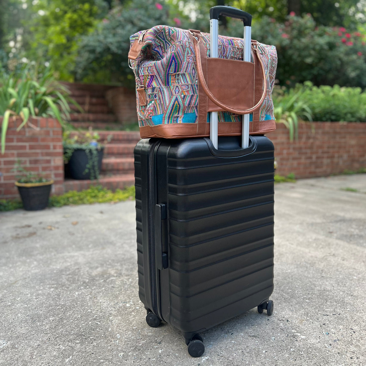 A multicolored geometric-patterned overnight travel bag with a brown leather base and a black trolley sleeve, placed on the ground next to a suitcase.