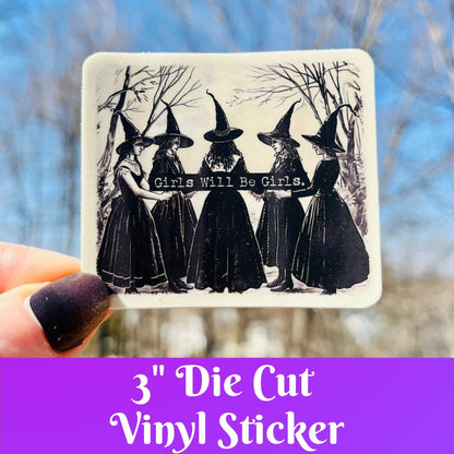 A hand holds a square die-cut vinyl sticker featuring a black-and-white illustration of five witches in pointed hats with the text &
