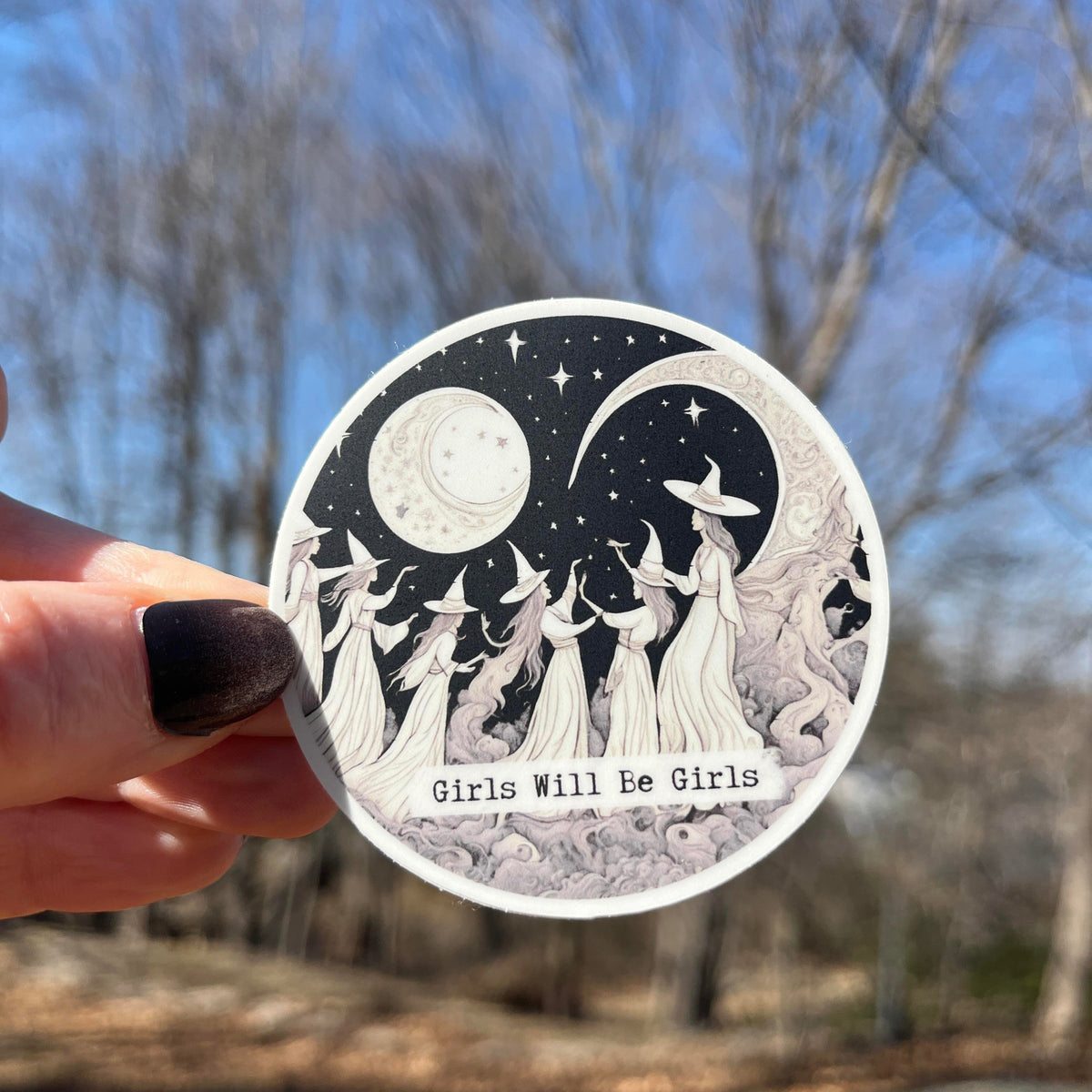 A hand with dark nail polish holds a round sticker featuring a black and white illustration of girls in witch hats under a crescent moon and stars, with the text &
