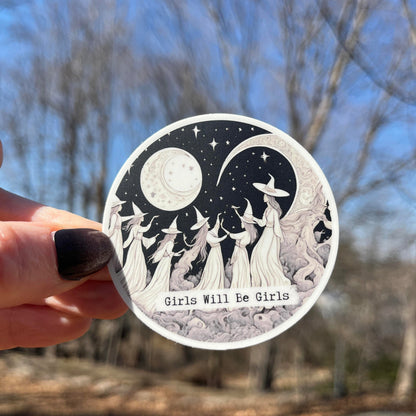 A hand with dark nail polish holds a round sticker featuring a black and white illustration of girls in witch hats under a crescent moon and stars, with the text &