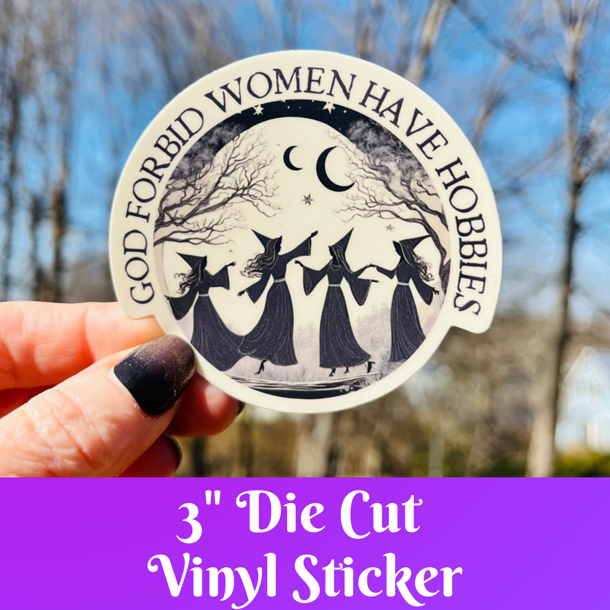 A hand holds a round die-cut vinyl sticker featuring a black and white illustration of three witches dancing under a crescent moon, with the text &
