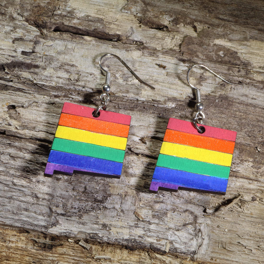 Hand-painted New Mexico state wooden dangle earrings featuring the LGBTQ+ Pride rainbow flag on hypoallergenic .925 sterling silver hooks. Lightweight Pride jewelry celebrating New Mexico, LGBTQ+ pride, equality, inclusion, and self-expression.