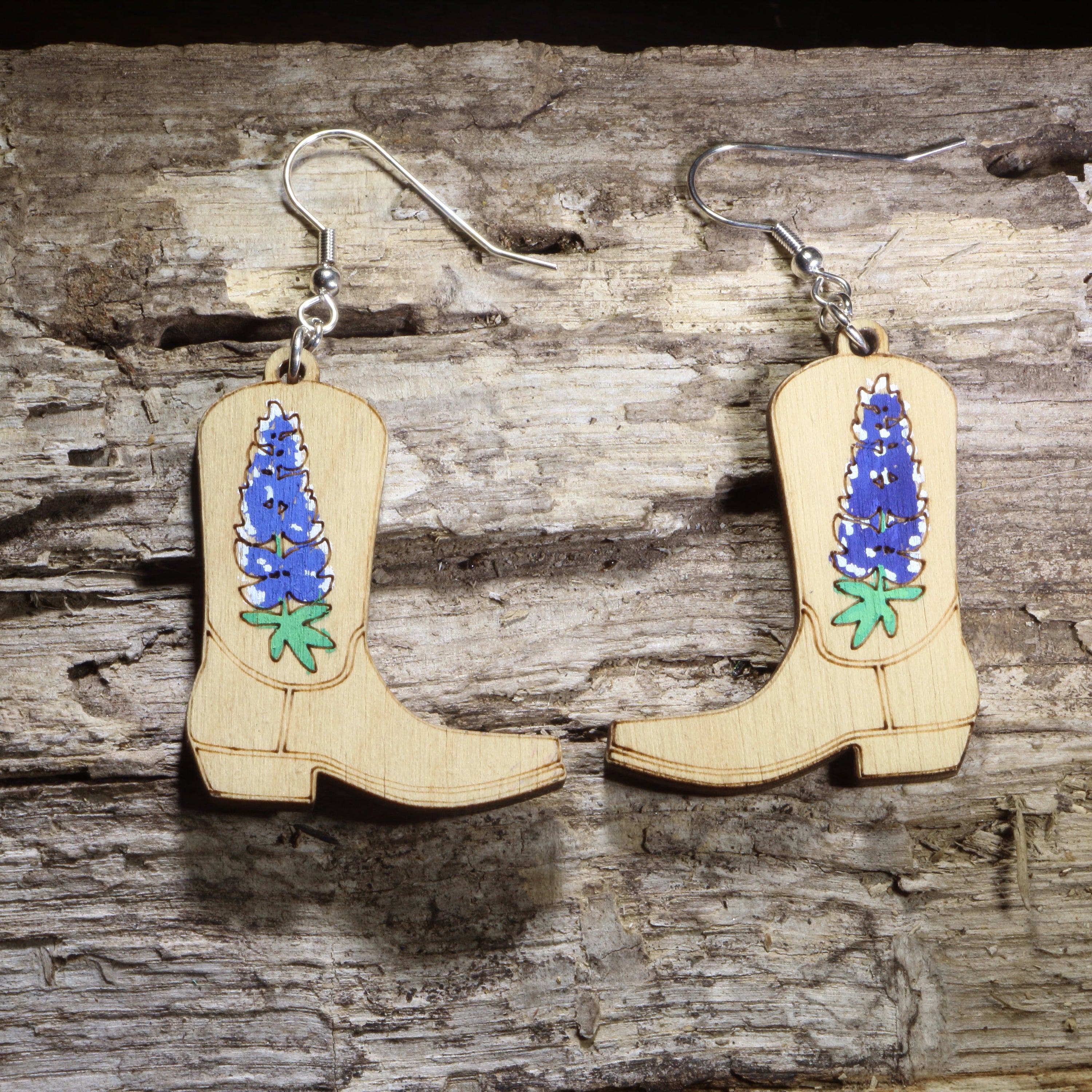 Hand-painted wooden cowboy boot dangle earrings featuring bluebonnet flowers on hypoallergenic .925 sterling silver hooks. Lightweight western floral jewelry for cowgirl style, bluebonnet lovers, rodeo outfits, country fashion, and Texas wildflower enthusiasts.