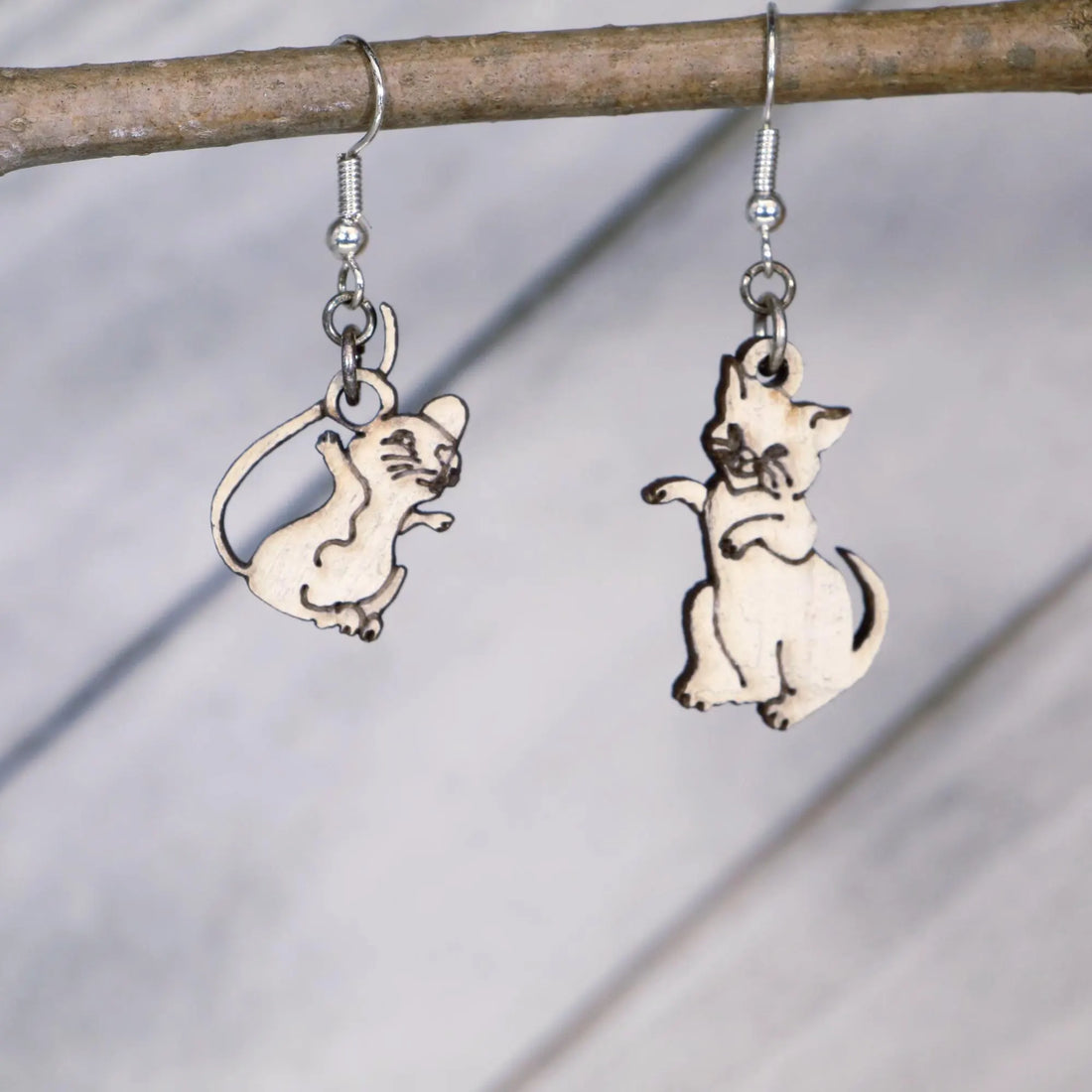Hand-painted wooden mismatched cat and mouse dangle earrings featuring a playful white cat reaching toward a smiling mouse on hypoallergenic .925 sterling silver hooks. Lightweight animal jewelry displayed hanging from a branch for cat lovers, mouse enthusiasts, pet owners, and fans of whimsical accessories.