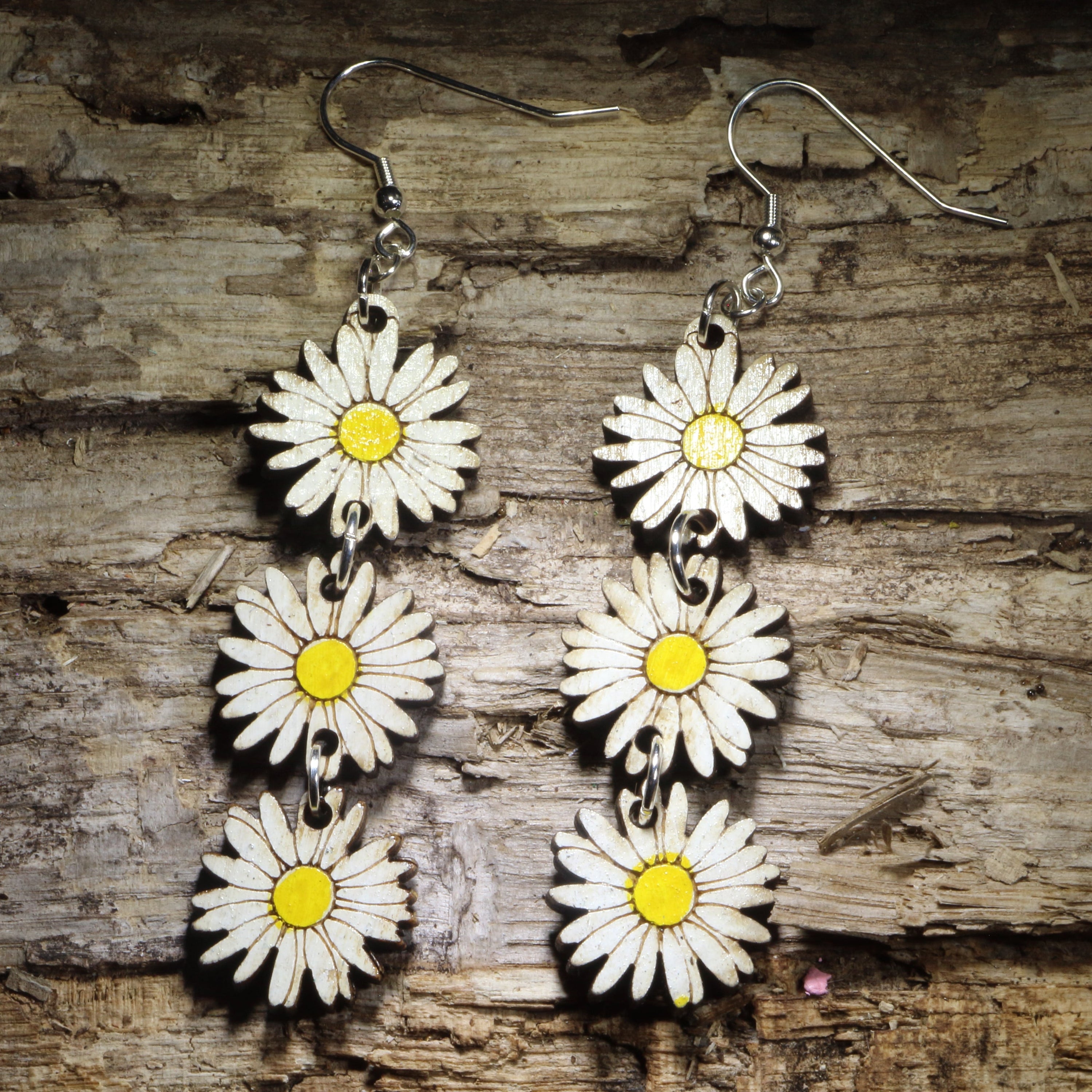 Handcrafted daisy chain wooden dangle earrings featuring three hand-painted white daisies with bright yellow centers linked together for graceful movement. Lightweight laser-cut floral earrings with hypoallergenic .925 sterling silver hooks, perfect for cottagecore fashion, wildflower lovers, spring and summer outfits, botanical jewelry, garden enthusiasts, and nature-inspired accessories