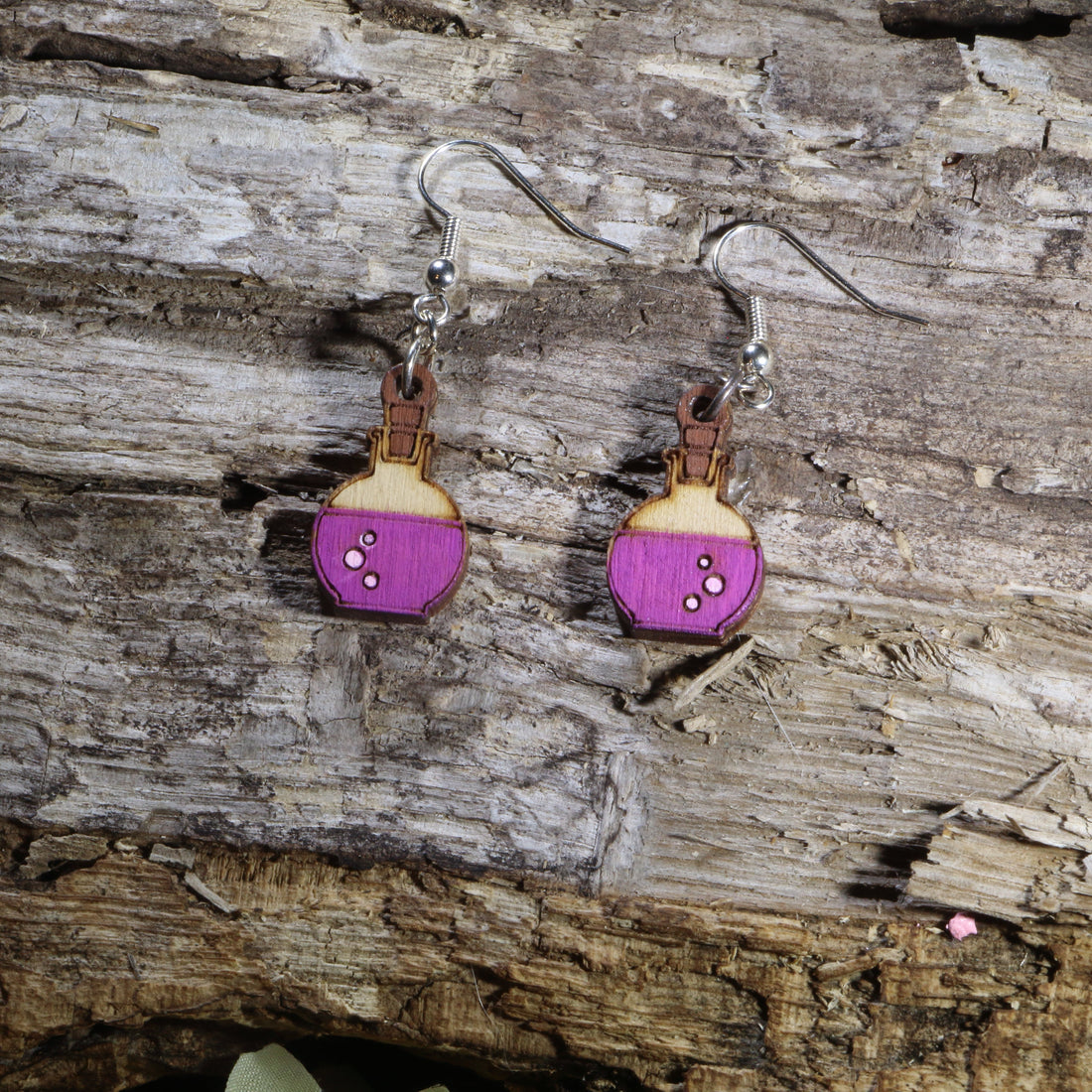 Handcrafted wooden potion bottle dangle earrings featuring vibrant purple potion details and engraved accents. These lightweight fantasy-inspired earrings are finished with hypoallergenic .925 sterling silver hooks, making them the perfect accessory for gamers, fantasy fans, witches, alchemists, tabletop RPG players, and lovers of magical jewelry.